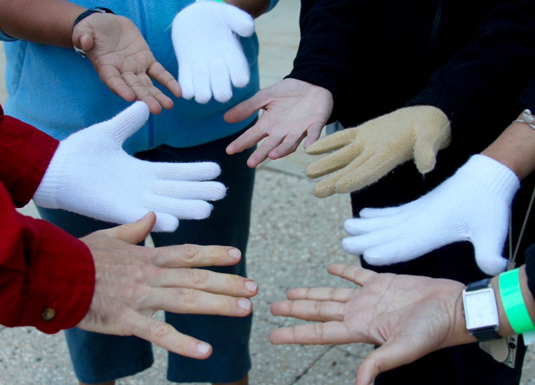 Photo showing eight hands with varied skin tones: 4 with gloves, 4 without. By sharing three sets of “emergency gloves” among the five of us, we found a way to share in the warmth: each of us with one hand covered, one uncovered. Together, we created some warmth, much laughter, and good memories that remind me still today that we need solutions for solidarity and mutual support. Together, we can create warmth, even in chilly conditions.