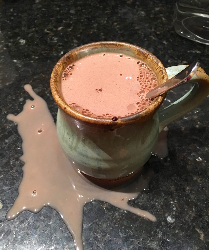 A green mug with a brown brim and handle contains hot chocolate and a spoon. Some of the hot chocolate has spilled onto the black marble countertop.