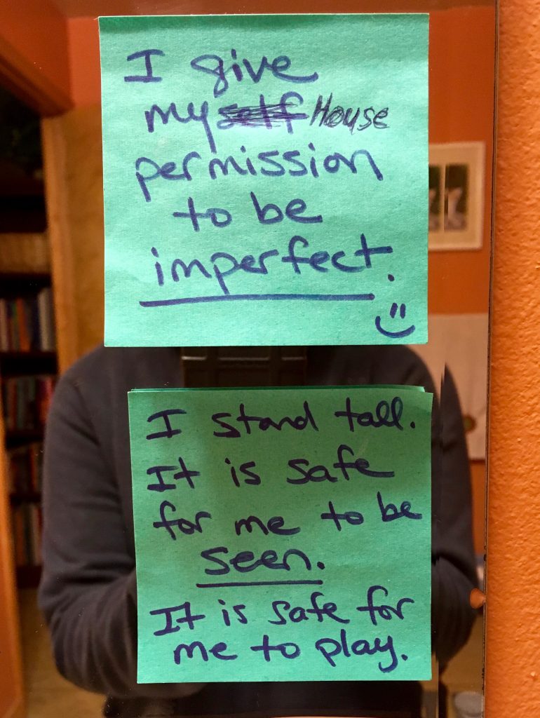 A mirror on an orange wall has two blue sticky-notes attached to it. On the first sticky-note reads, "I give my house permission to be imperfect." and the word "imperfect" is underlined. There is a small happy face next to it. The sticky-note below that reads, "I stand tall. It is safe for me to be seen. It is safe for me to play." where the word "seen" is underlined. The writing is done in a dark blue marker. In the reflection of the mirror is the upperboddy of a person wearing a grey sweater.