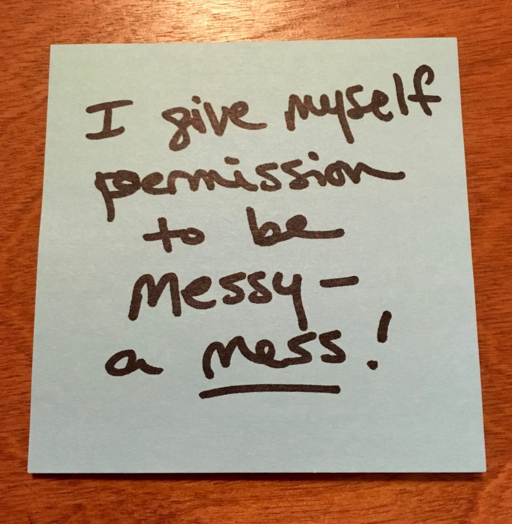 A blue sticky-note on an orange-brown wooden table reads, in black ink, "I give myself permission to be messy - a mess!" The word "mess" is underlined.