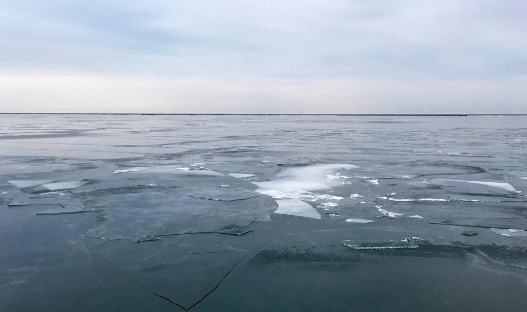 The sky is grey and cloudy. There is a large body of water, which is covered in a thin layer of fracturing ice. In places where the ice has broken, the water appears more white than the dark blue color of the rest of the water.