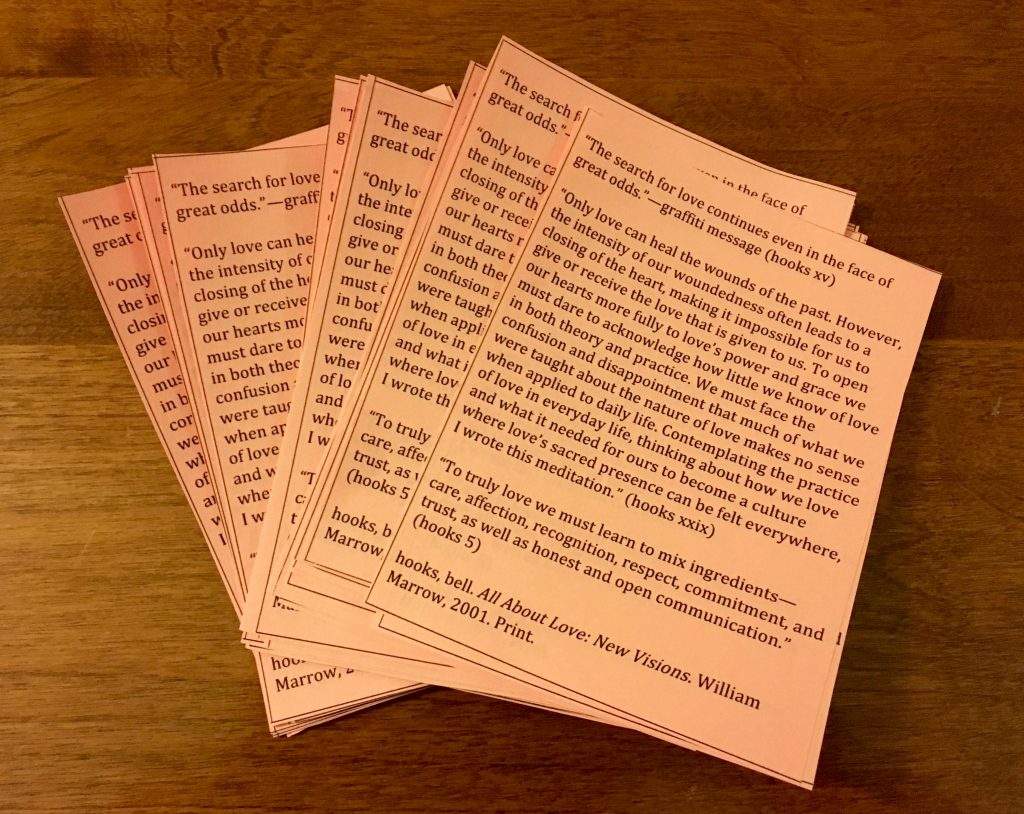 An image of a stack of peach orange colored papers with balck print on them on top of a light brown wooden table. The stack of papers is slightly fanned out. The print on the papers is an excerpt from bell hooks' "All About Love: New Visions."