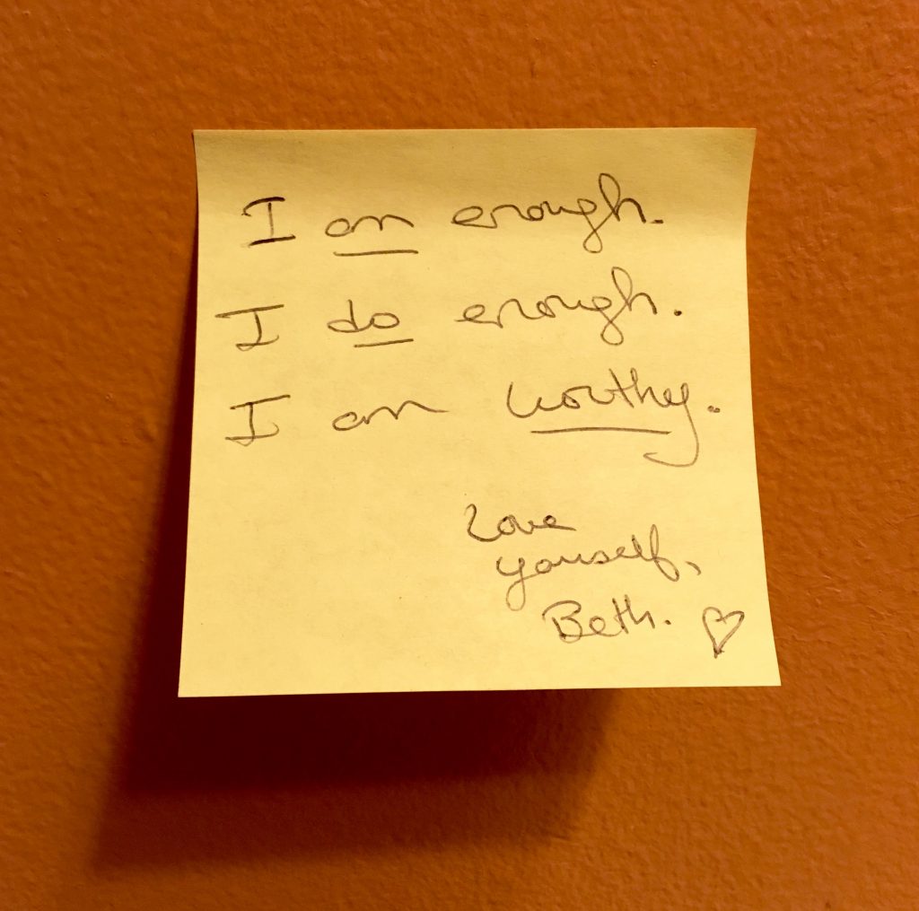 An image of a yellow sticky note on an orange wall. The note reads in black pen, "I am enough. I do enough. I am worthy. Love yourself, Beth." The words "am," "do," and "worthy" are underlined. Next to "Beth." is a small doodle of a heart.