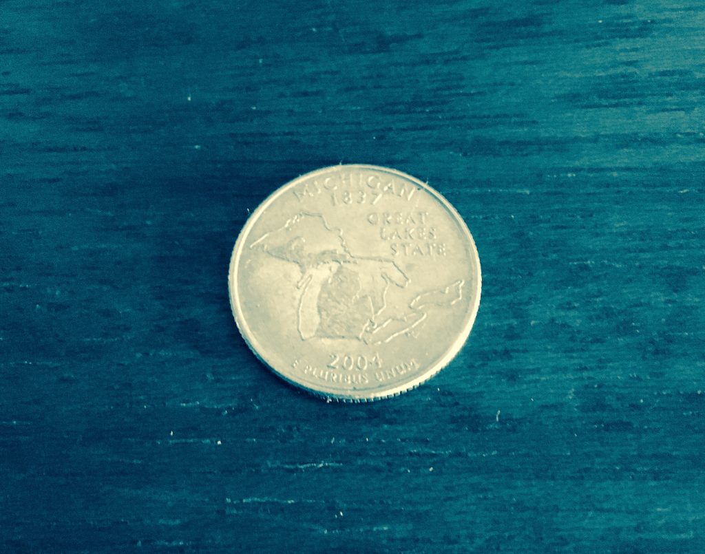 A Michigan quarter on a blue wooden surface. The quarter reads "Michigan, Great Lake State, 1837, 2004," and is engraved with an image of the state of Michigan.