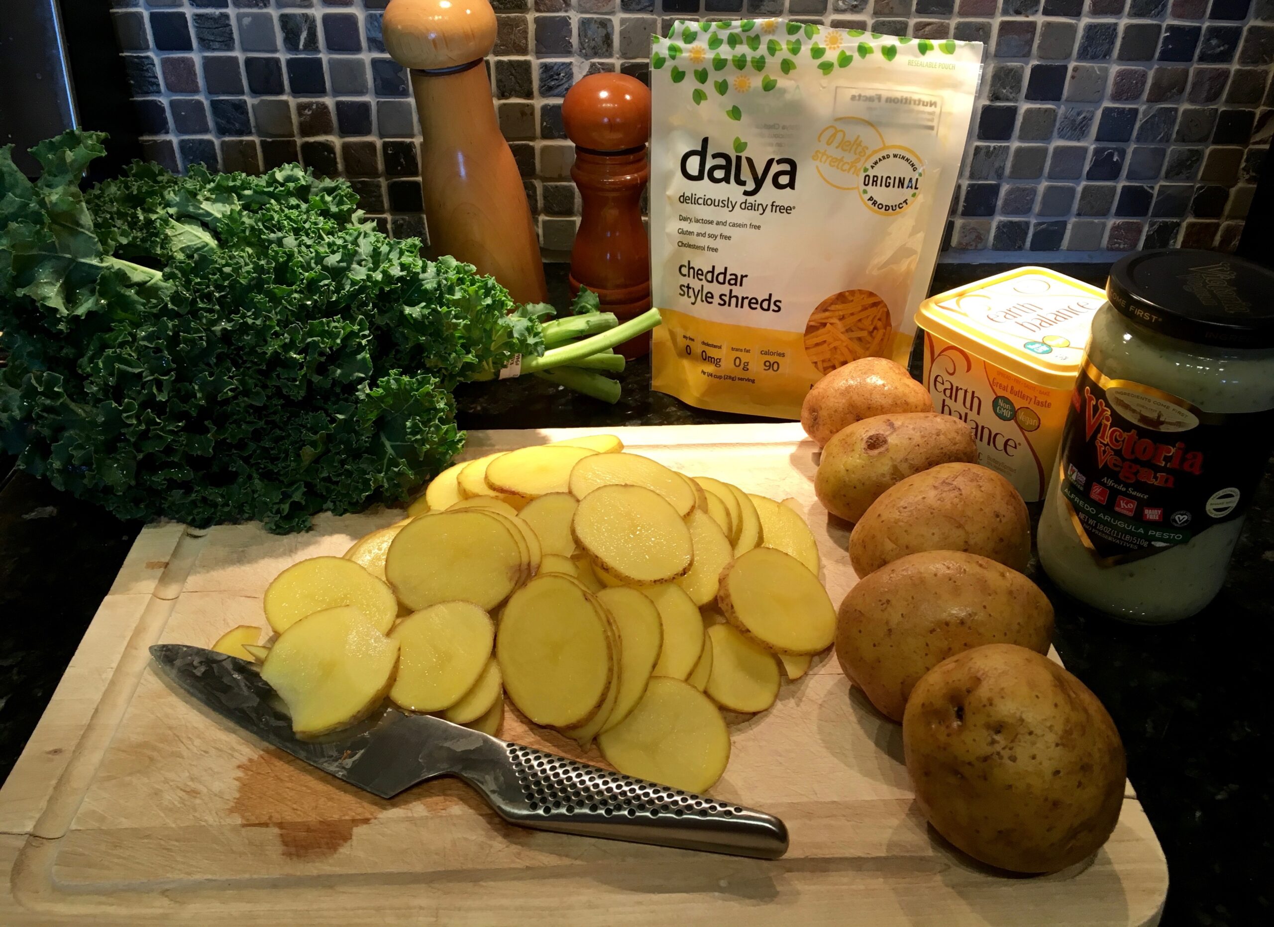 An image of an assortment of ingredients. Sliced potatoes on a cutting board next to whole potatoes, a jar of Victoria Vegan pesto alfredo sauce, a container of Earth Balance fake butter, Daiya dairy free chedar cheese, salt and pepper grinders, and a buschel of kale.