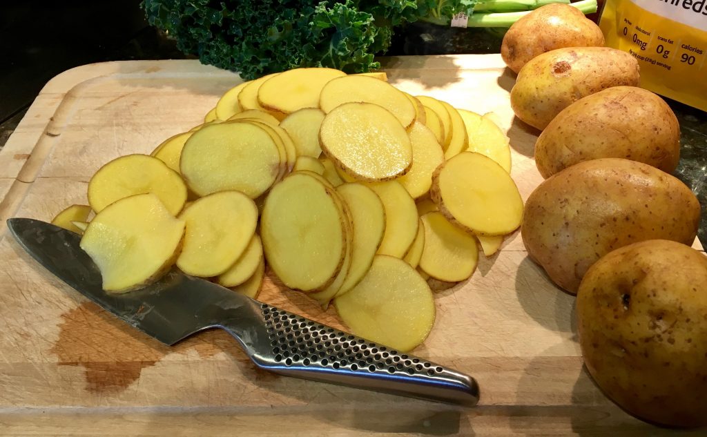 An image of sliced potatoes on a cutting board with a metal knife. On the side of the cutting board is 5 whole potatoes.