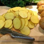 An image of sliced potatoes on a cutting board with a metal knife. On the side of the cutting board is 5 whole potatoes.
