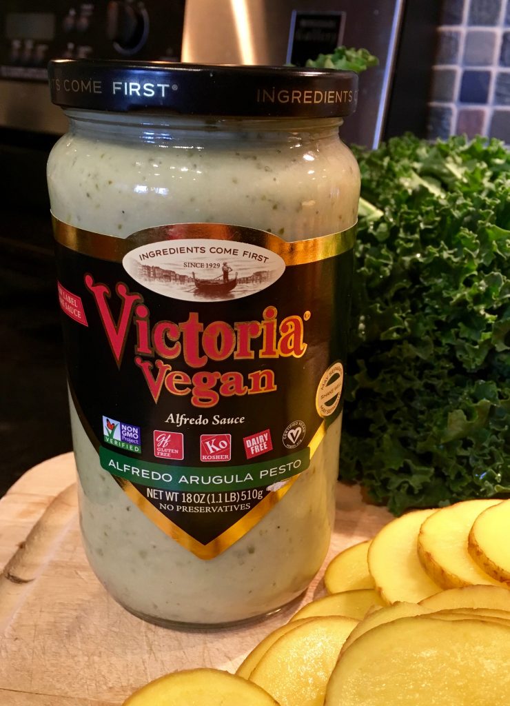 A bottle of Victoria Vegan alfredo arugula pesto next to thinly sliced potatoes and in front of kale.