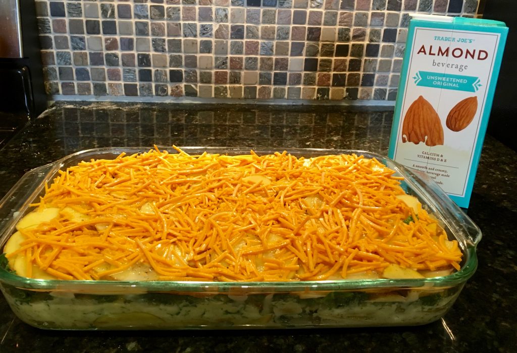 An image of a dairy free cheese covered potato and kale casserole in a glass dish on a marble countertop. Next to the dish is a teal and white carton of Trader Joe's almond beverage.