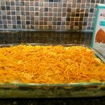 An image of a dairy free cheese covered potato and kale casserole in a glass dish on a marble countertop. Next to the dish is a teal and white carton of Trader Joe's almond beverage.