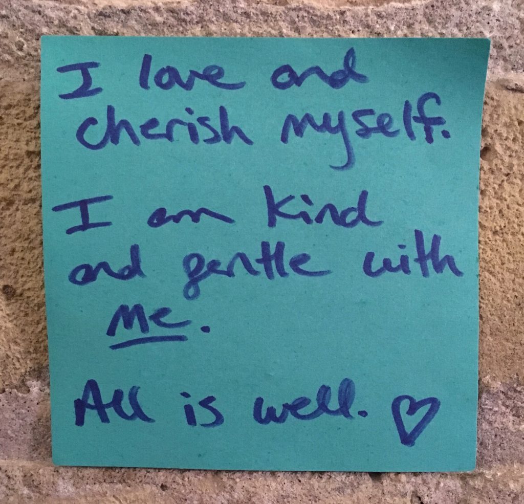 A blue sticky note with dark blue writing that reads "I love and cherish myself. I am kind and gentle with me. All is well." A small doodle of a heart appears next to "well" and "me" is underlined.