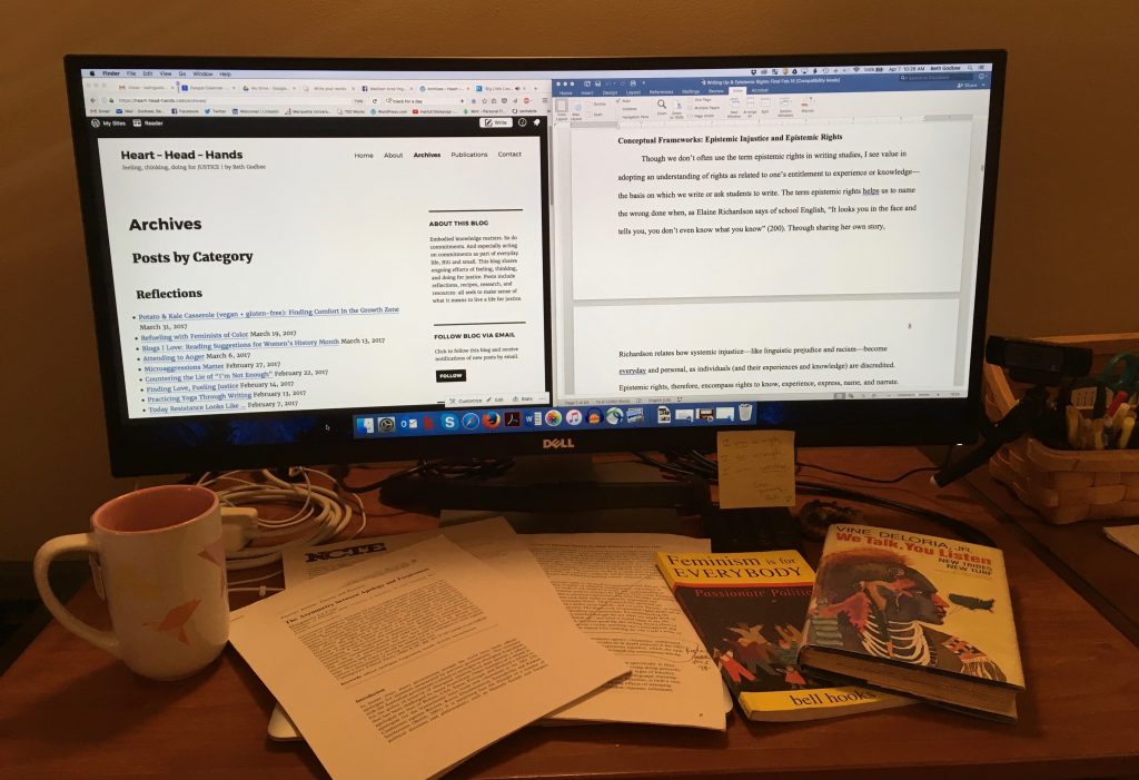An image of a desk with a large computer monitor, a pink mug, papers spread across the keyboard, and two books. The books are "Feminism is for EVERYBODY, Passionate Politics," by bell hooks, and "We Talk, You Listen," by Vine Deloria Jr. On the computer screens is an open Word document and a tab showing the archives of Heart-Head-Hands.com.