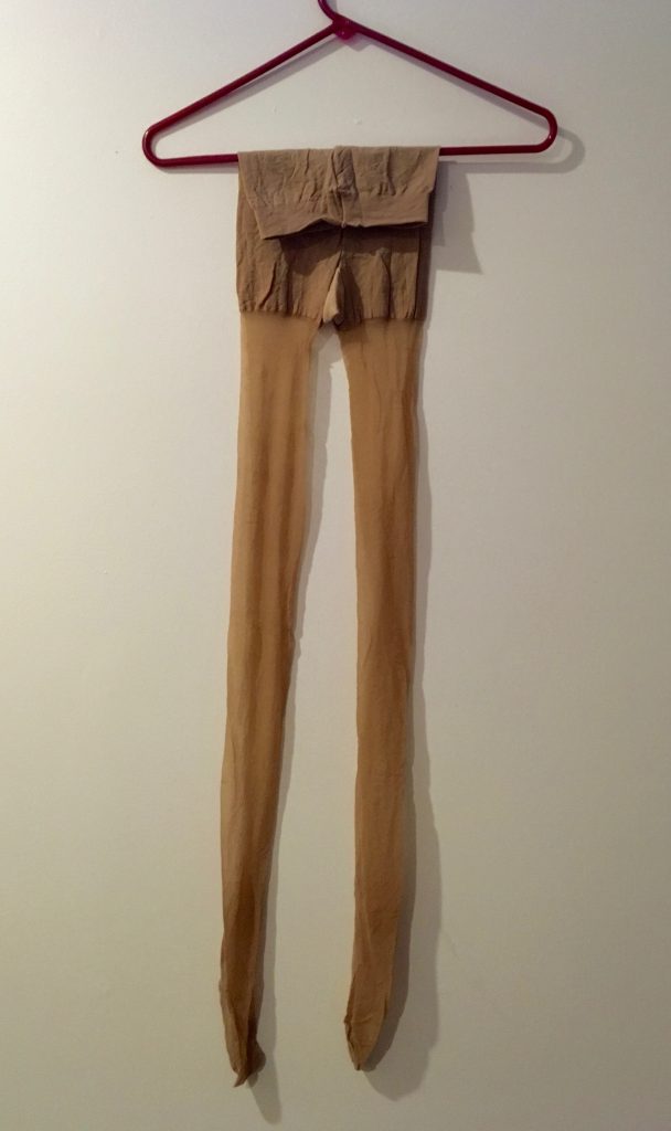 Tan stockings hang on a red coat hanger on a white wall.