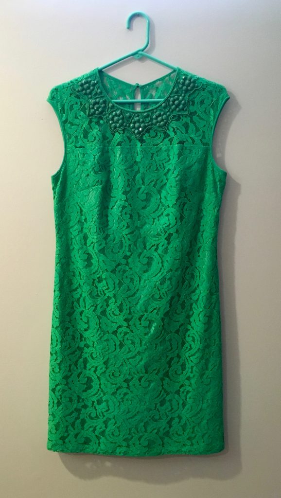 A lacy green dress on a green coathanger against a white wall. The top of the dress is embroidered with green jewels.