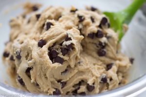 Chocolate chip cookie dough being mixed in a glass bowl with a green spoon.