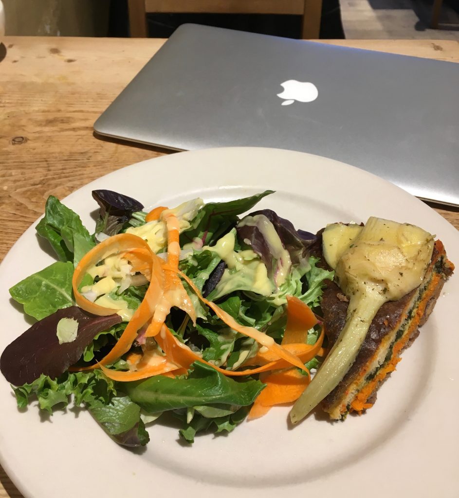 A plate of salad and a slice of quiche in front of a silver laptop. The items are on a wood grain table.