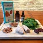 Ingreidents for a smoothie sit in a white wooden tray on top of a wooden table. The ingredients include a carton of almond beverage, a scoop of cacao powder, almonds, frozen banana, dates, a bottle of liquid stevia, and a bottle of vanilla extract.