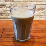 A beige-light green smothie sits in a gass on a wooden table. In the background of the photo is a white brick wall.