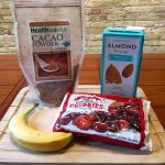 Smoothie ingredients sit on a white wooden tray on a orange-brown wooden table. The ingredients include a bag of cacao powder, a carton of almond beverage, a banana, and a frozen bag of cherries.