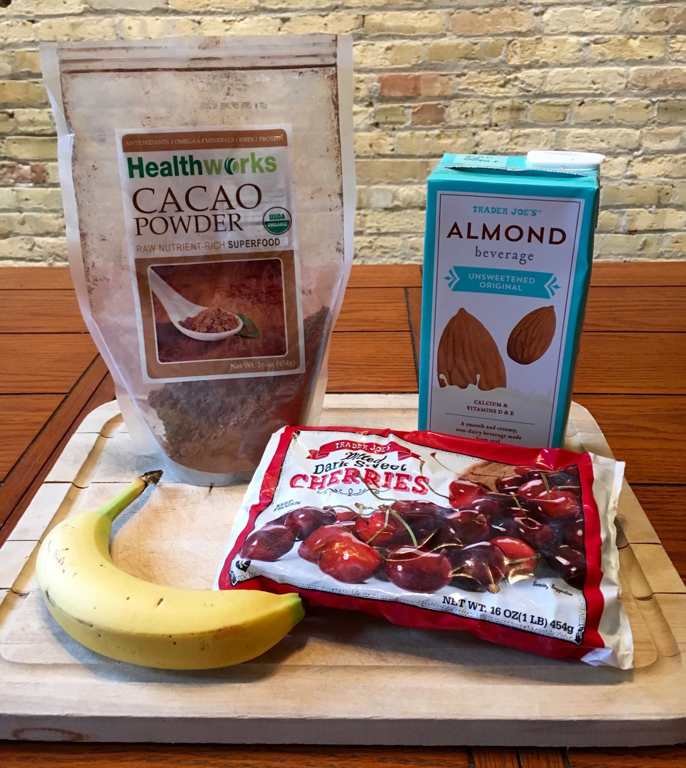 Smoothie ingredients sit on a white wooden tray on a orange-brown wooden table. The ingredients include a bag of cacao powder, a carton of almond beverage, a banana, and a frozen bag of cherries.