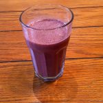 A red-purple smoothie with a bit of froth on top is in a glass on a wooden table.