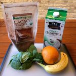 Ingredients for a smoothie sit on a counter. The ingredients include a bag of cacao powder, a carton of organic coconut milk, a handful of spinach, an orange, and a banana.