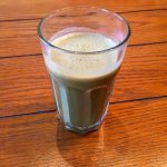 A glass is filled with a beige smoothie mixture with a bit of froth on the top. The glass sits on top of a wooden table.