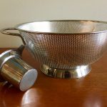 A silver colander and smaller strainer, which lays on it's side, sit on a brown wooden table.