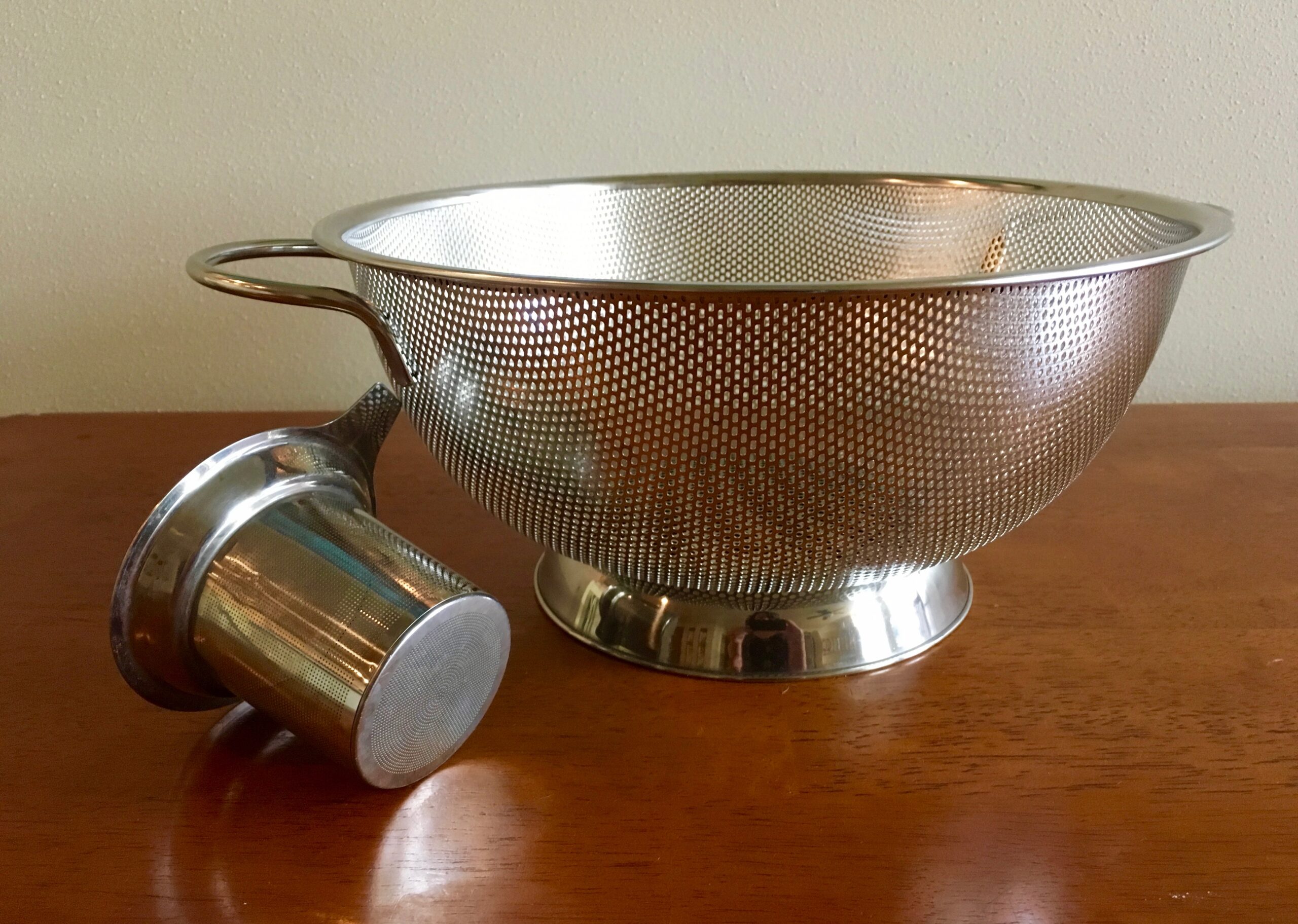 A silver colander and smaller strainer, which lays on it's side, sit on a brown wooden table.