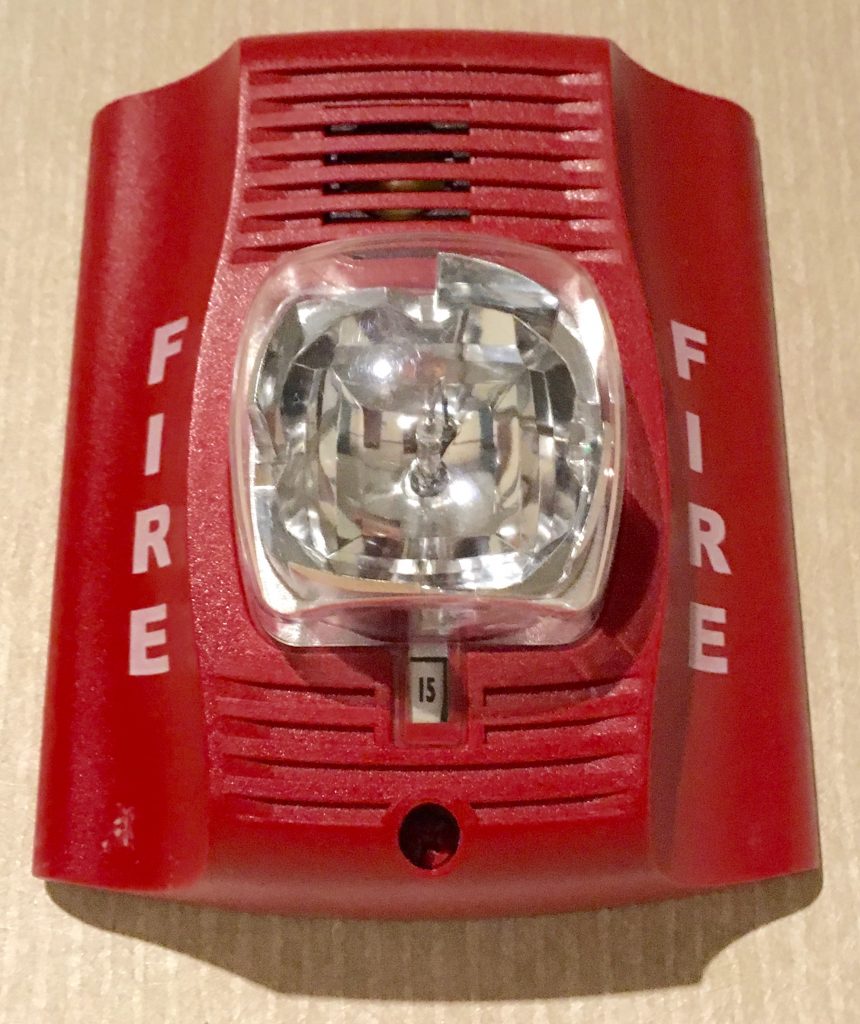A red, plastic fire alarm with a clear light in the middle and "FIRE" written vertically in white on both sides.