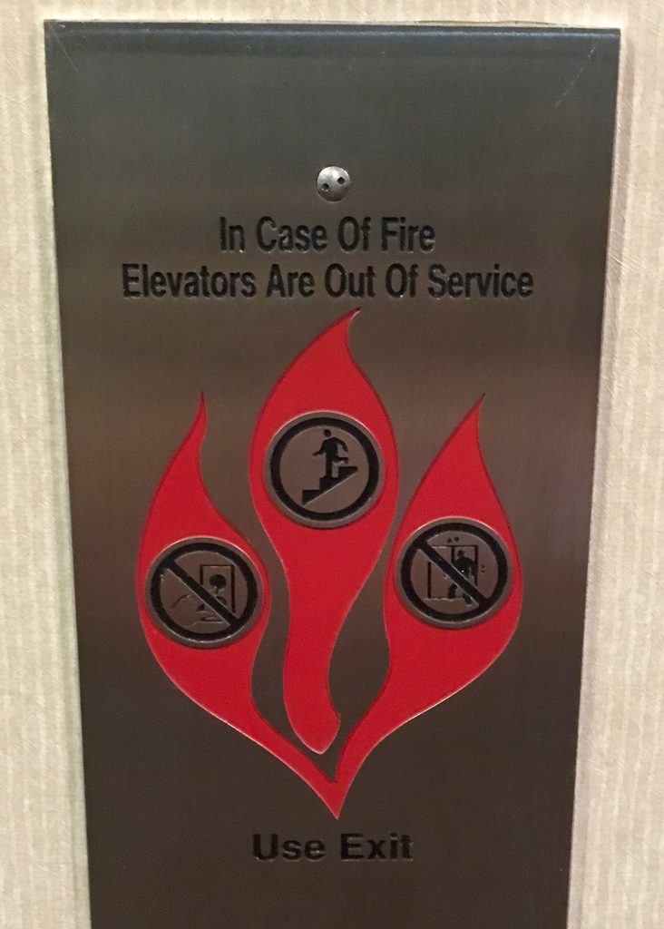 A silver fire information pannel is screwed into a wall. It reads "In Case of Fire, Elevators Are Out Of Service, Use Exit." Below the writing is a drawing of a flame with three prongs. The one on the left shows to not press the elevator bell, the middle one depicts using the stairs, and the right image depicts not entering the elevator.