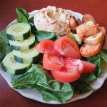 A small white plate on a reddish-brown table. On the plate is a bed of spinach, a few cucumber slices, tomato slices, hummus, and croutons.