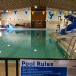 This image shows an indoor swimming pool with the YMCA logo against the back wall, a blue water slide to the right, and a sign with "Pool Rules" posted in the front.