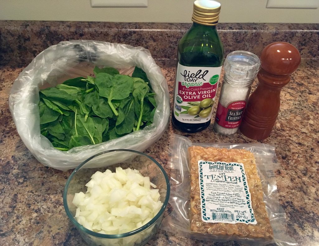 An array of ingredients needed to cook sauteed spinach and tempeh. The ingredients include a bag of spinach, a bowl of chopped onions, a package of tempeh, a bottle of olive oil, and salt and pepper. The ingredients sit on a brown marble counter.