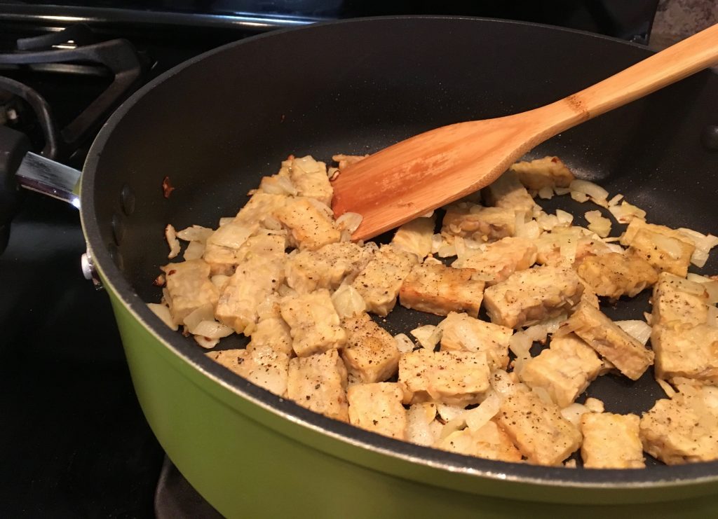 Tempeh sits in a green pan with a wooden spatula as it begins to cook.