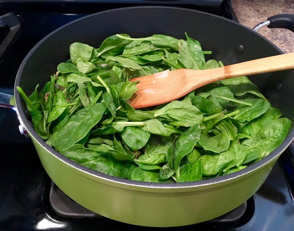 Spinach sits in a green pot with a wooden spatula sticking out as it begins to cook.