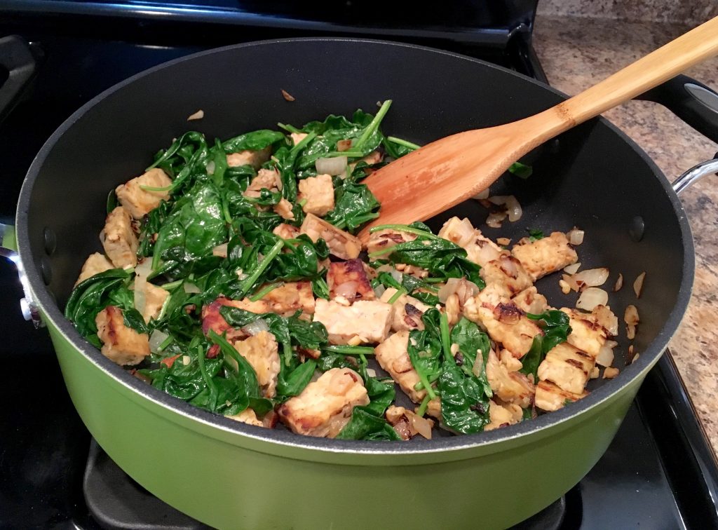 A green pot with a wooden spatula sticking out. In the pot is spinach that is being sauteed and tempeh.
