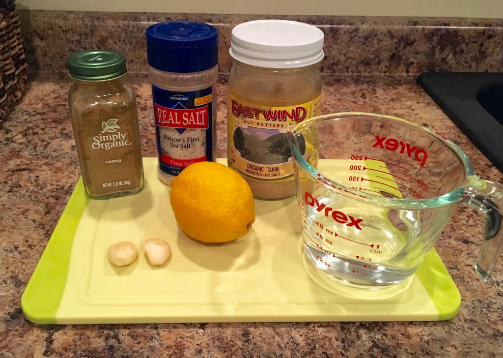 The ingredients needed to make tahini suace sit on a yellow, plastic cutting board on a brown countertop. The ingredients include a measuring cup with water, a lemon, two garlic cloves, cumin powder, sea salt, and a container of organic tahini.