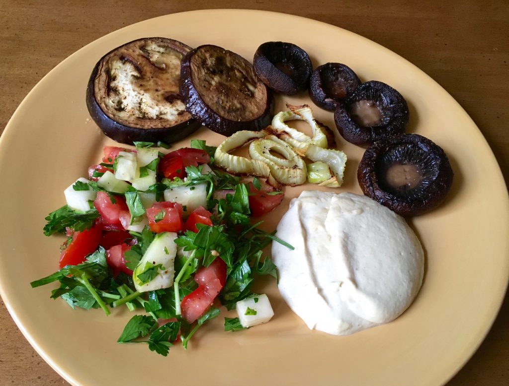 A fresh veggie salad, roasted mushrooms and onions, and tahini sauce on a beige plate on a wooden surface.