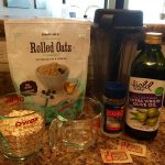 A group of ingredients sit on a counter top including olive oil in a bottle, a blender, a bag of rolled oats, a measuring cup filled with oats, a measuring cup containing water, a bottle of salt, and two sugar packets. These ingredients are for a pie crust.