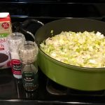 A green pot containing uncooked vegetables sits on a stove next to a container of vegetable broth and smaller containers of salt and pepper.