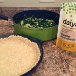 The ingredients needed for a savory, vegan pie sit on a countertop. A blue dish is lined with pie crust, a green pot which contains a cooked green vegetable medley, and a bag of dairy free cheddar cheese.