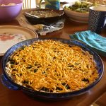 A savory, fake cheese covered pie in a blue clear dish sits on a crowded dinner table that has all sorts of other dishes on it.