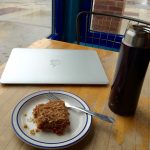 A silver laptop, a brown water bottle, and a white plate with blue edges topped with a baked treat and fork sit on a wooden table in a cafe.