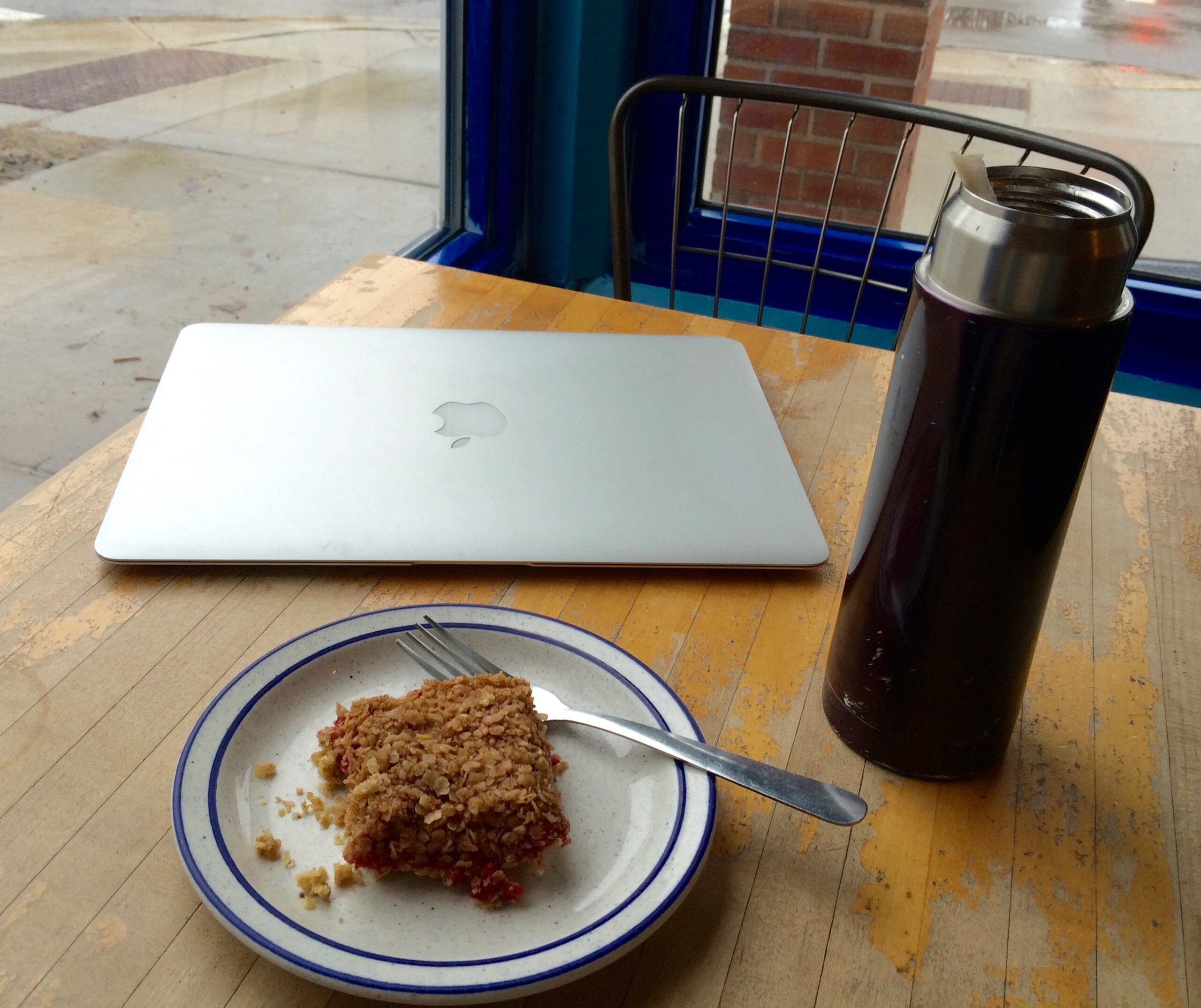 A silver laptop, a brown water bottle, and a white plate with blue edges topped with a baked treat and fork sit on a wooden table in a cafe.