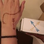A brown temporary tatoo of a snake and arrow on Beth's hand which is also adorned by a brown, gem bracelet. In Beth's other hand she holds a white box with a blue arrow and yellow arrow stamped on the side.