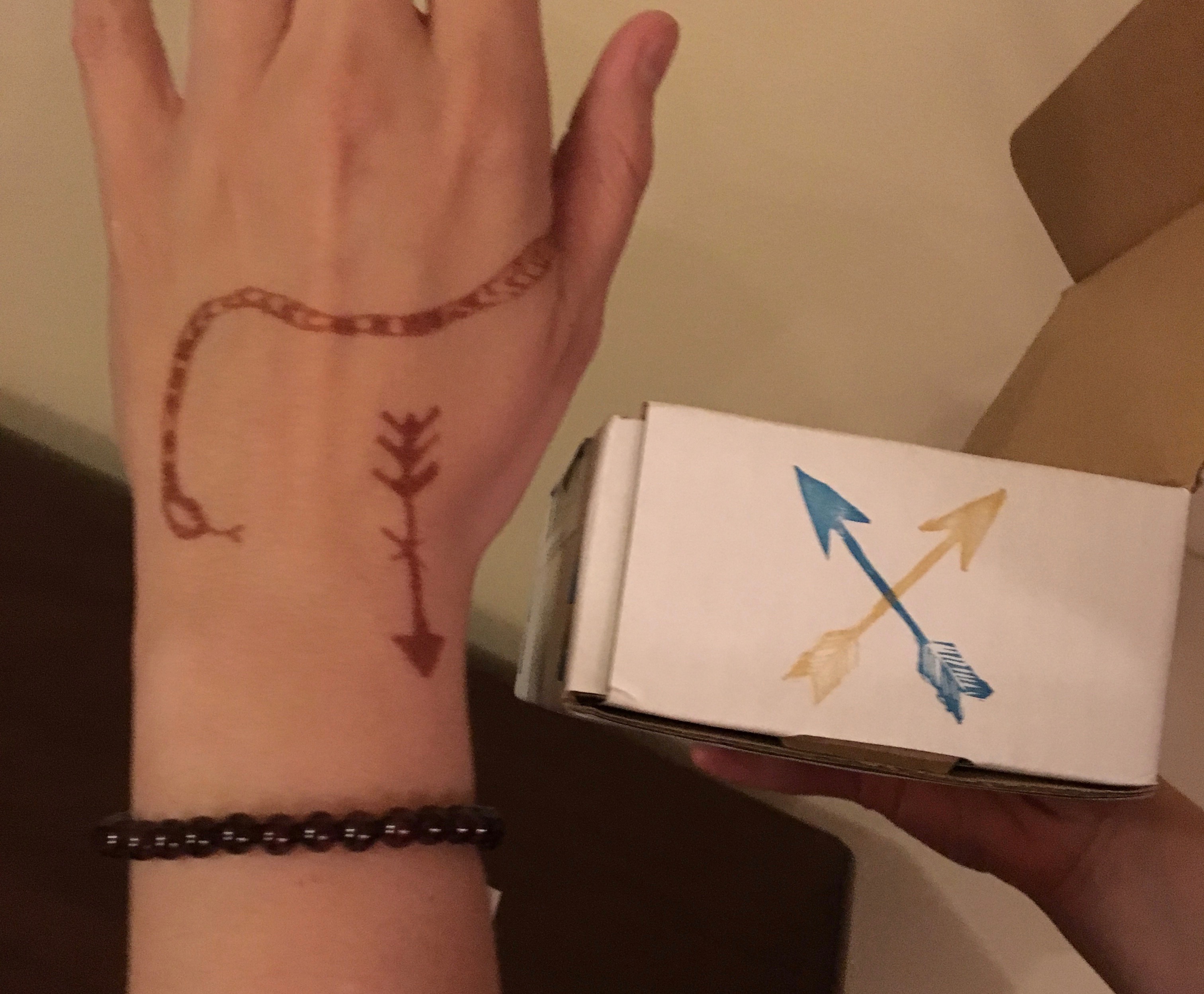 A brown temporary tatoo of a snake and arrow on Beth's hand which is also adorned by a brown, gem bracelet. In Beth's other hand she holds a white box with a blue arrow and yellow arrow stamped on the side.