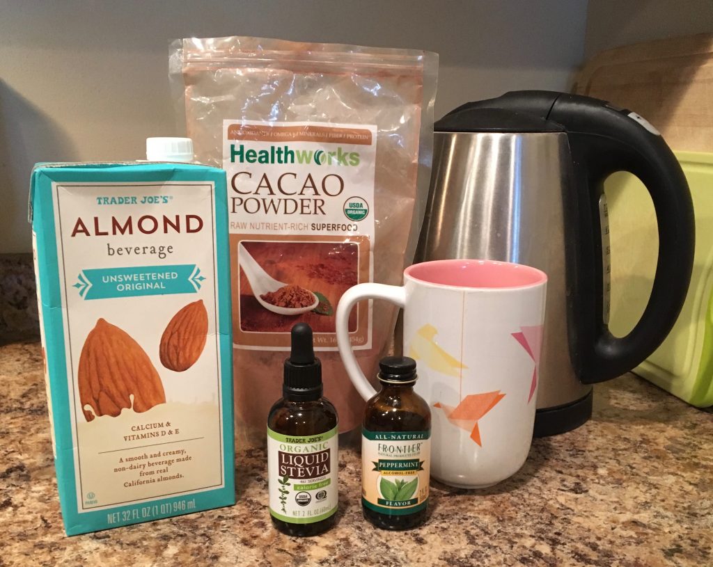 The ingredients for homemade hot chocolate are setup on a brown marble countertop. The ingredients include a hot water heater, a pink and white mug, a bag of cacao powder, a carton of almond beverage, a bottle of liquid stevia, and a small bottle of peppermint.