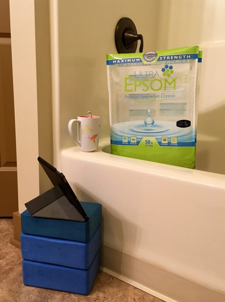 This photo shows a large bag of Epsom salt sitting in a bathtub, along with a mug and iPad set up for watching shows.