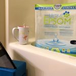 A cozy bath setup with a tub containing a large bag of epsom salt crystals with a tablet propped up on yoga blocks outside of it. On the rim of the tub is a pink and white mug.